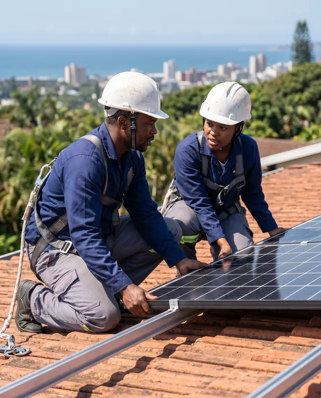 Two installers in hard hats fitting a solar panel on a tiled roof, with Durban and the sea behind them